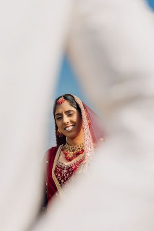 Creative portrait of bride in red wedding attire and jewelry through white fabric frame.