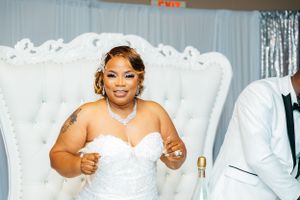 Bride in white dress poses elegantly on ornate white throne at wedding reception.