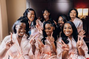 A group of bridesmaids in pink robes showing peace signs and laughing together.