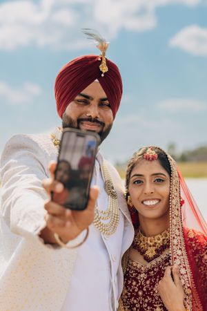 A couple in traditional wedding attire takes a selfie together outdoors.