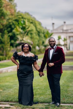 Elegant couple in formal attire poses together on mansion grounds with lush greenery in background.