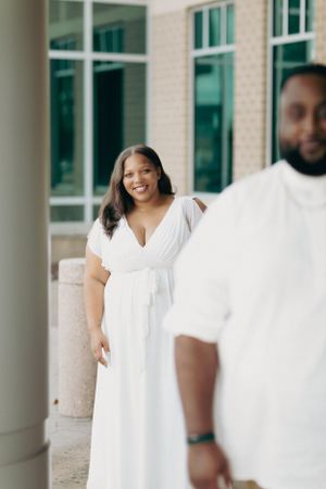 A couple in white outfits poses outside a modern building with glass windows.