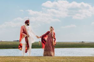 A couple in traditional Sikh wedding attire stands by a lake holding hands.