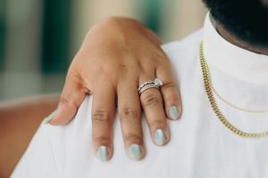 Close-up shot of a hand wearing an engagement ring and wedding band against a white shirt with gold chain.