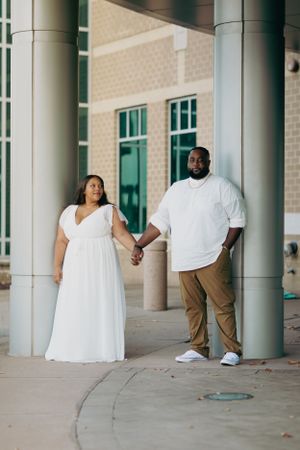 A couple in coordinated white outfits holds hands while standing between classical columns.