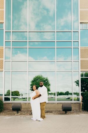 Couple poses in casual white outfits against modern glass building facade.