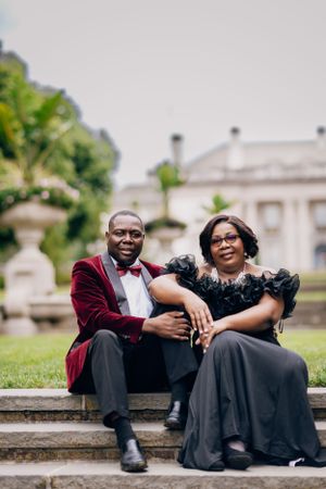 Couple in elegant evening wear sits together on stone steps of historic mansion.