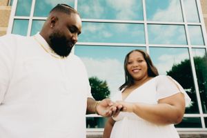 A couple in white clothing looks down at an engagement ring outdoors near a modern building.