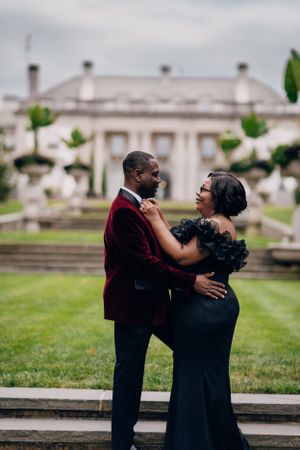 Couple in formal wear embraces on garden steps with grand mansion in background.