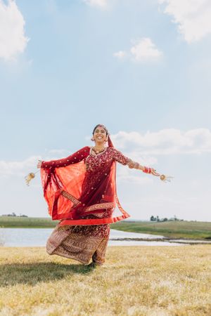A bride in a red embroidered lehenga twirls outdoors against a bright blue sky.