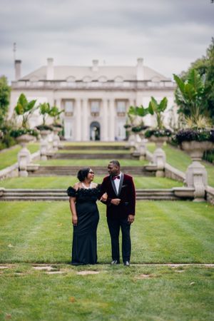 A couple shares a moment on manicured grounds with a grand white mansion in the background.