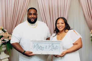 A couple in white outfits hold up a decorative 'Mr & Mrs' sign against a pink curtain backdrop with floral arrangements.