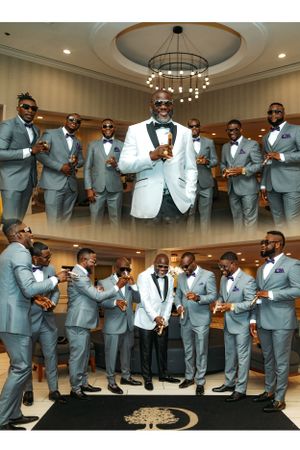 Wedding party poses in formal grey suits with groom in white tuxedo under elegant chandelier.