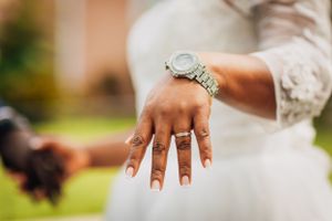 Close-up of an elegantly manicured hand displaying a silver wristwatch and wedding ring against a white dress.