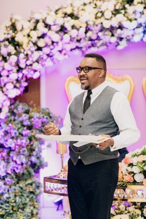 A speaker in a grey vest addresses guests at a reception with floral arch backdrop.