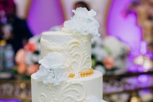 Elegant white wedding cake with intricate swirl patterns and floral decorations displayed against a purple backdrop.