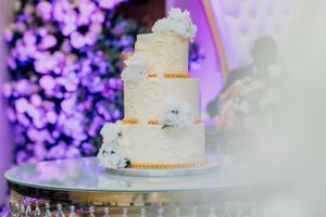 Three-tiered white wedding cake decorated with delicate patterns and flowers against purple lighting.