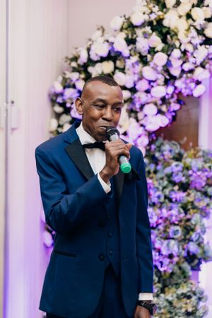 Speaker in navy blue tuxedo performing in front of a purple and white floral backdrop.