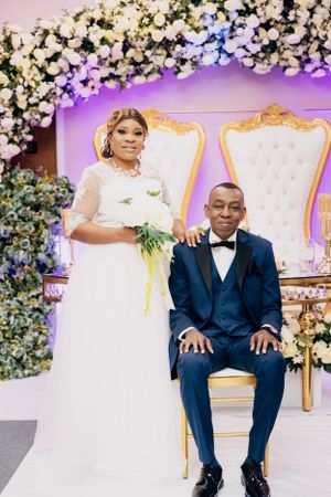 Wedding couple poses in front of floral backdrop with elegant white chairs and purple lighting.