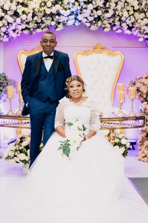 Formal wedding portrait with ornate floral backdrop and elegant white throne chairs.