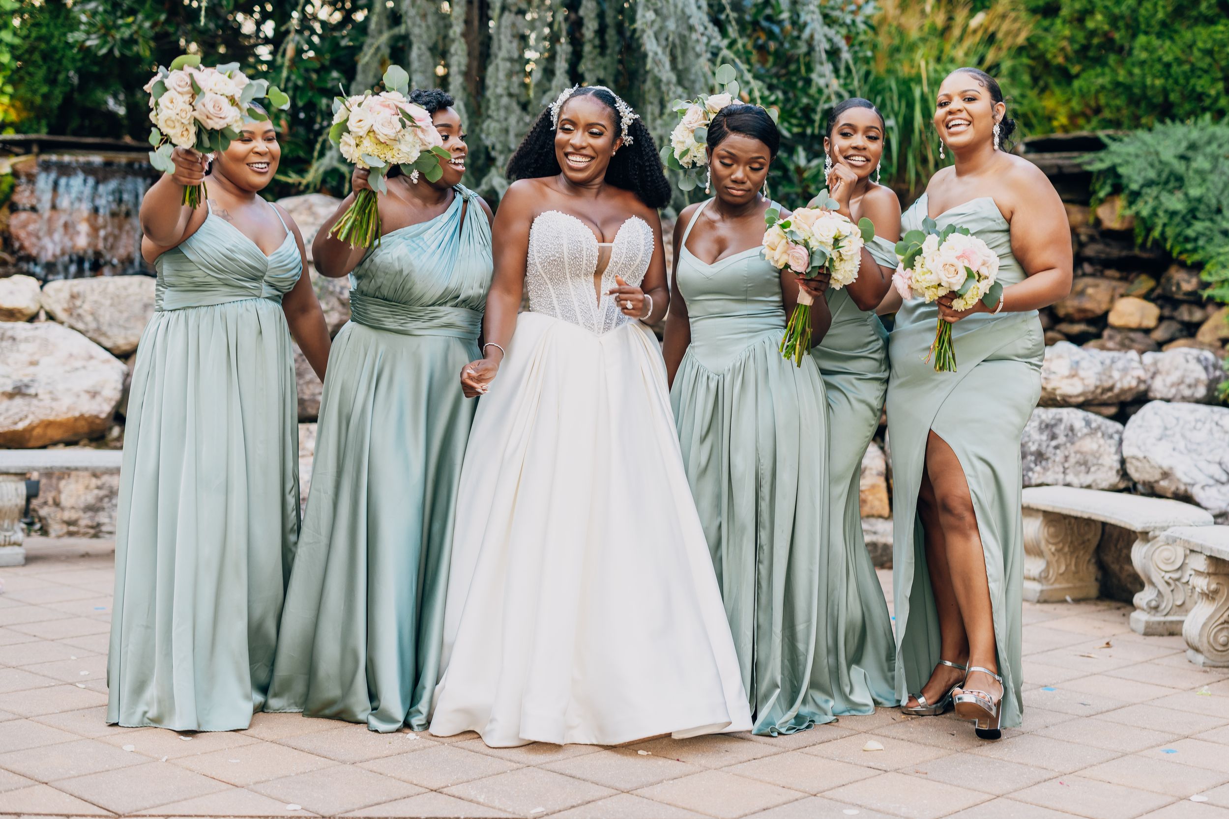 Bridal party in sage green dresses poses outdoors against stone wall.