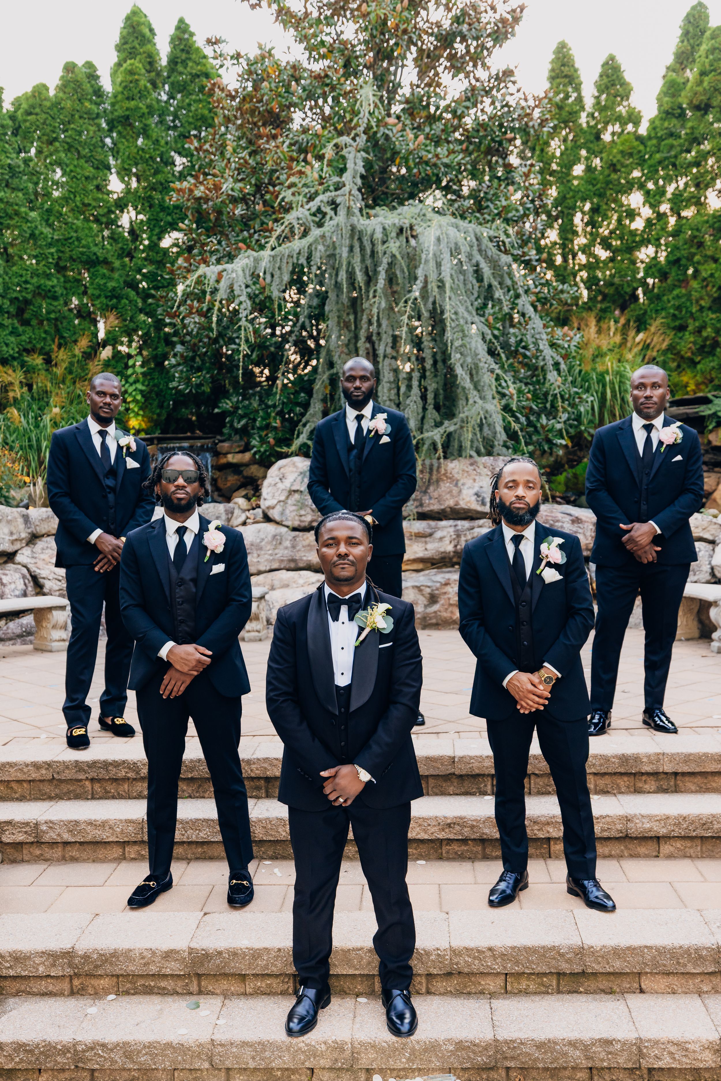 A wedding party dressed in black tuxedos standing on stone steps in an outdoor garden setting at sunset.