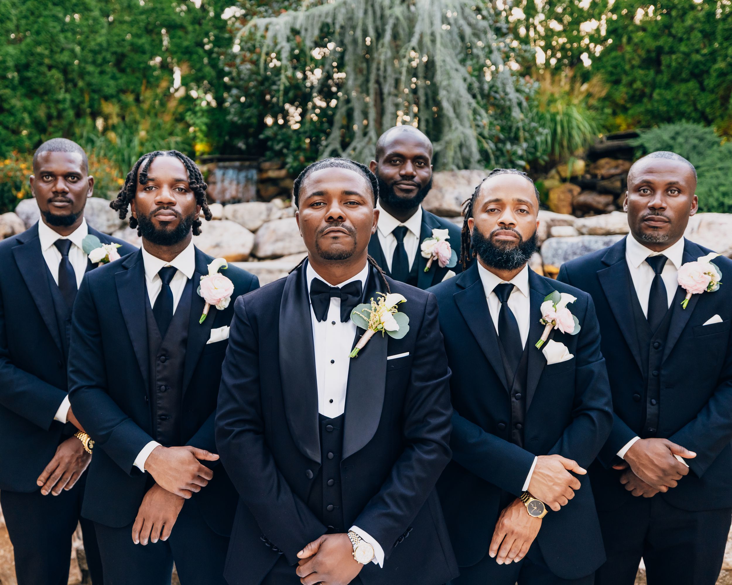Wedding party in classic black tuxedos and white boutonnieres pose outdoors.