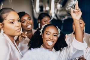 A group of bridesmaids in white robes share candid laughs during wedding preparation.