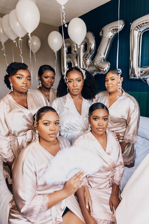 Bridesmaids in blush pink robes pose together during wedding preparations with white balloons.