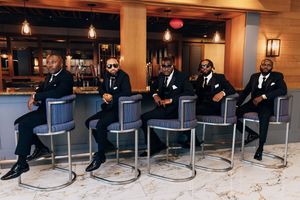 Groomsmen sitting in a row on modern barstools in formal attire.