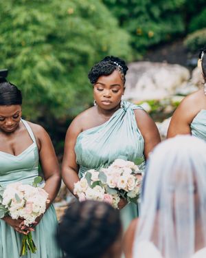 Wedding party in sage green dresses holding white floral bouquets gather for an outdoor ceremony.