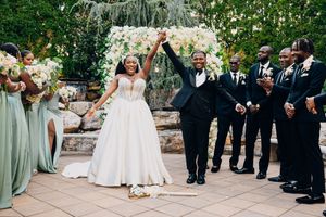 Wedding party members in sage green dresses and black suits gather on patio with stone columns and lush greenery.