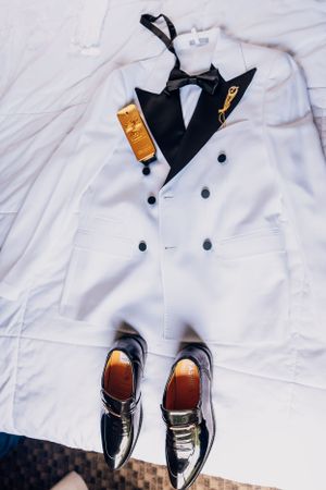 A white double-breasted tuxedo jacket with black bow tie and dress shoes laid flat on white fabric.