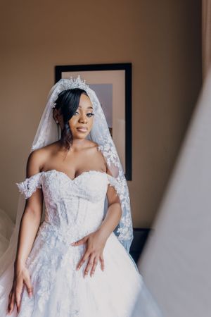 Bride in off-shoulder lace wedding gown and tiara poses elegantly before ceremony.