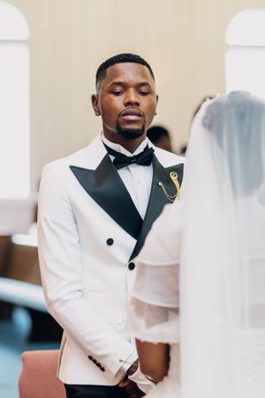 A close-up shot of a groom in a white tuxedo with black lapels and bowtie during a wedding ceremony.