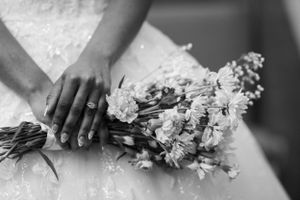 Black and white photo of hands holding delicate white flowers with a lace wedding dress in the background.