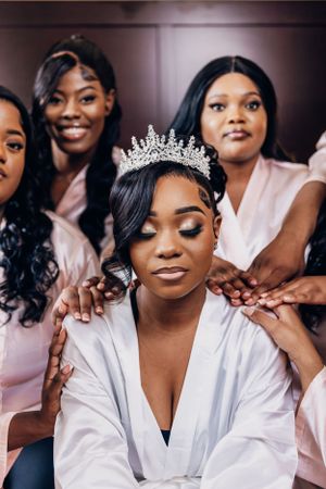 Bride wearing tiara poses with bridesmaids in pink robes during pre-wedding preparations.