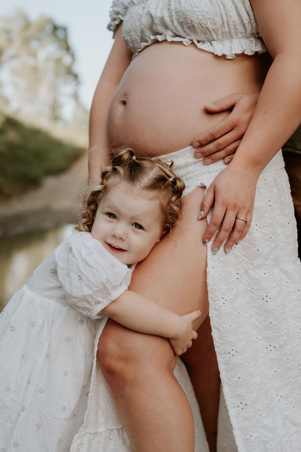 Toddler girl in white dress hugging pregnant mother's belly outdoors near water in a maternity photo.