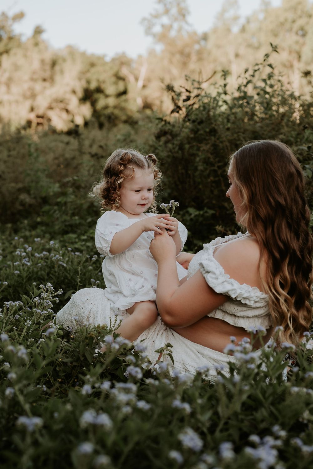A pregnant woman in a white dress sits in a wildflower field with her curly-haired toddler holding small flowers.