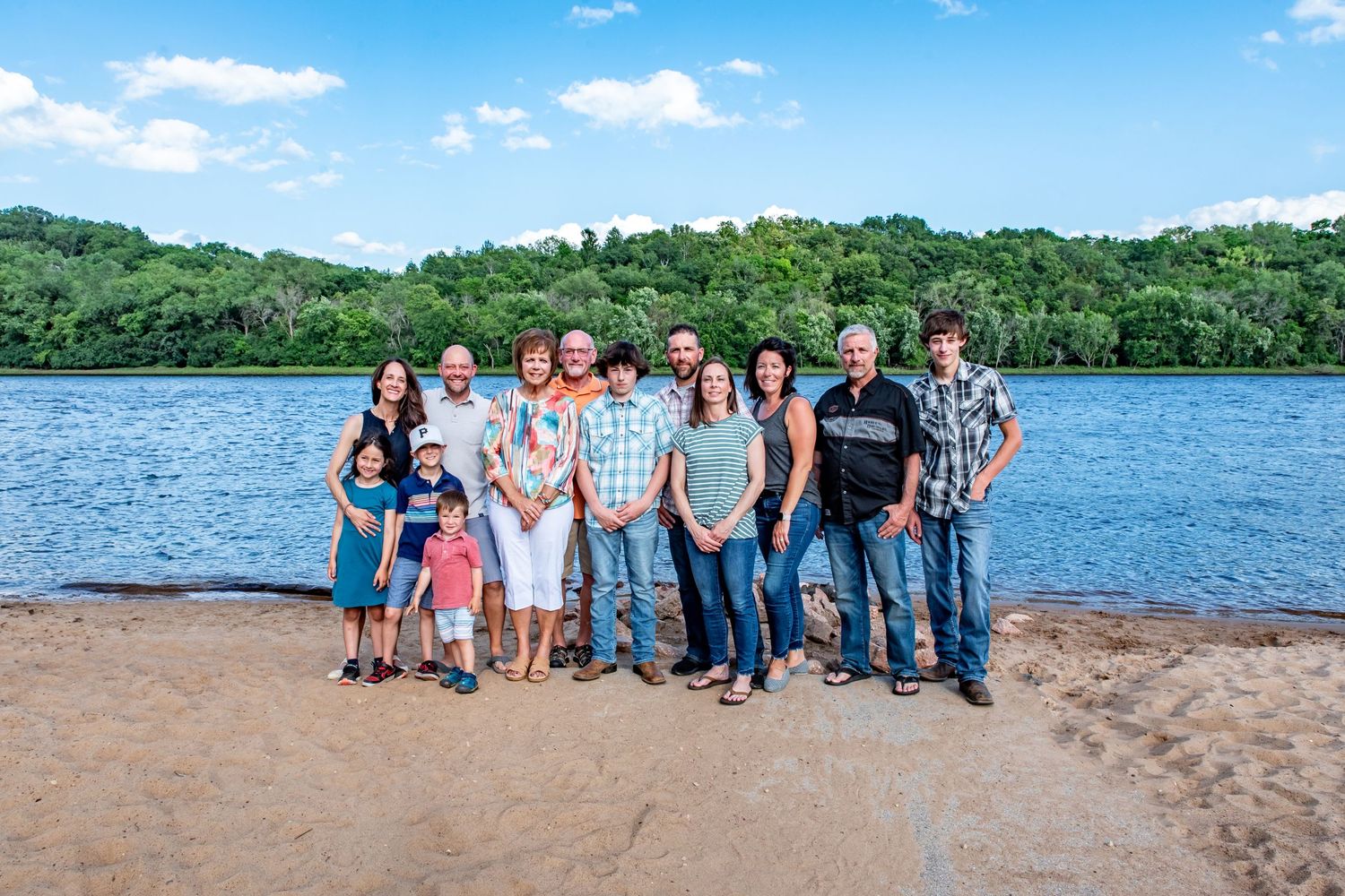 Large group gathered on sandy beach with lake and green hills in background.