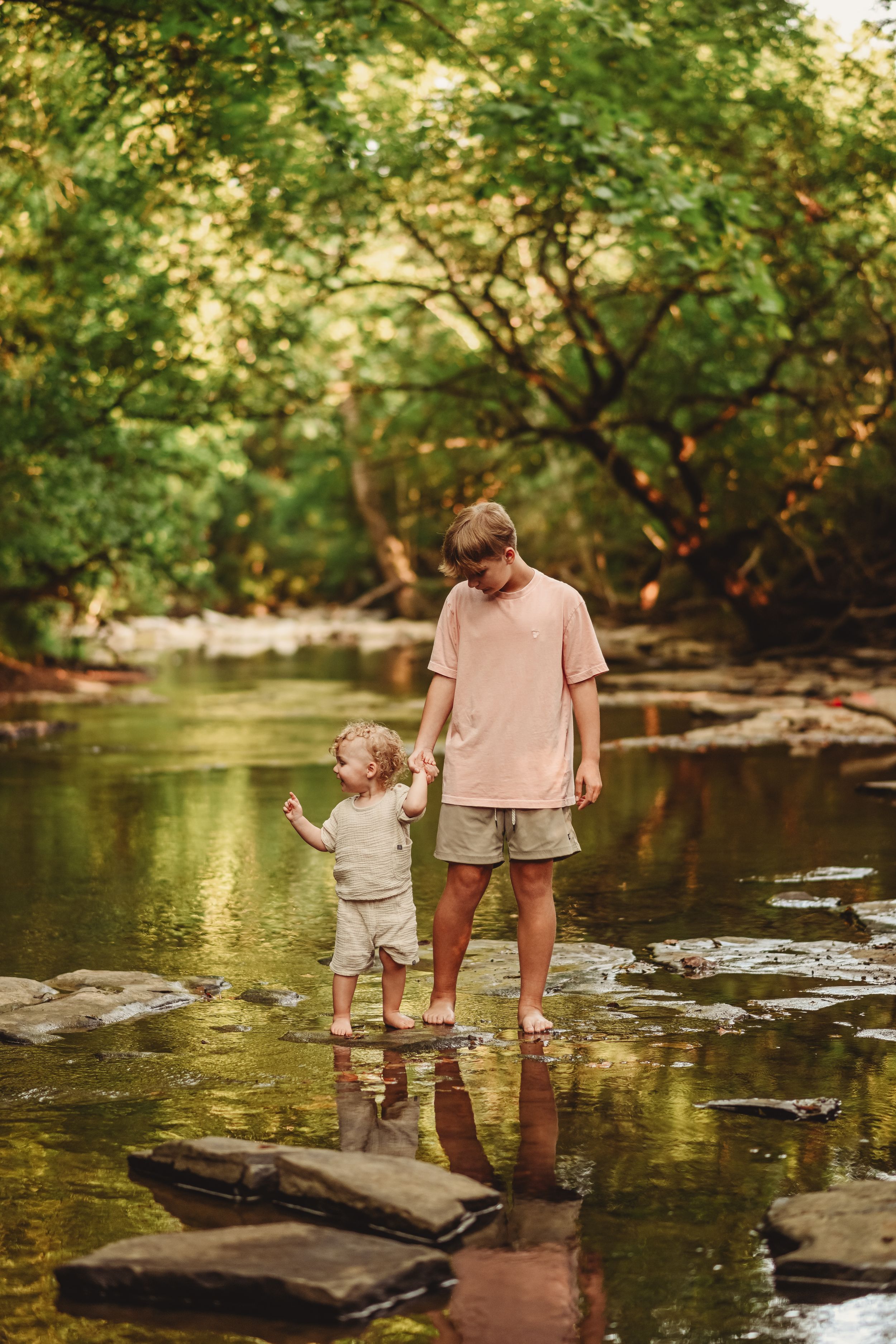 Two people stand on rocks in a peaceful creek with trees reflecting in the calm water.