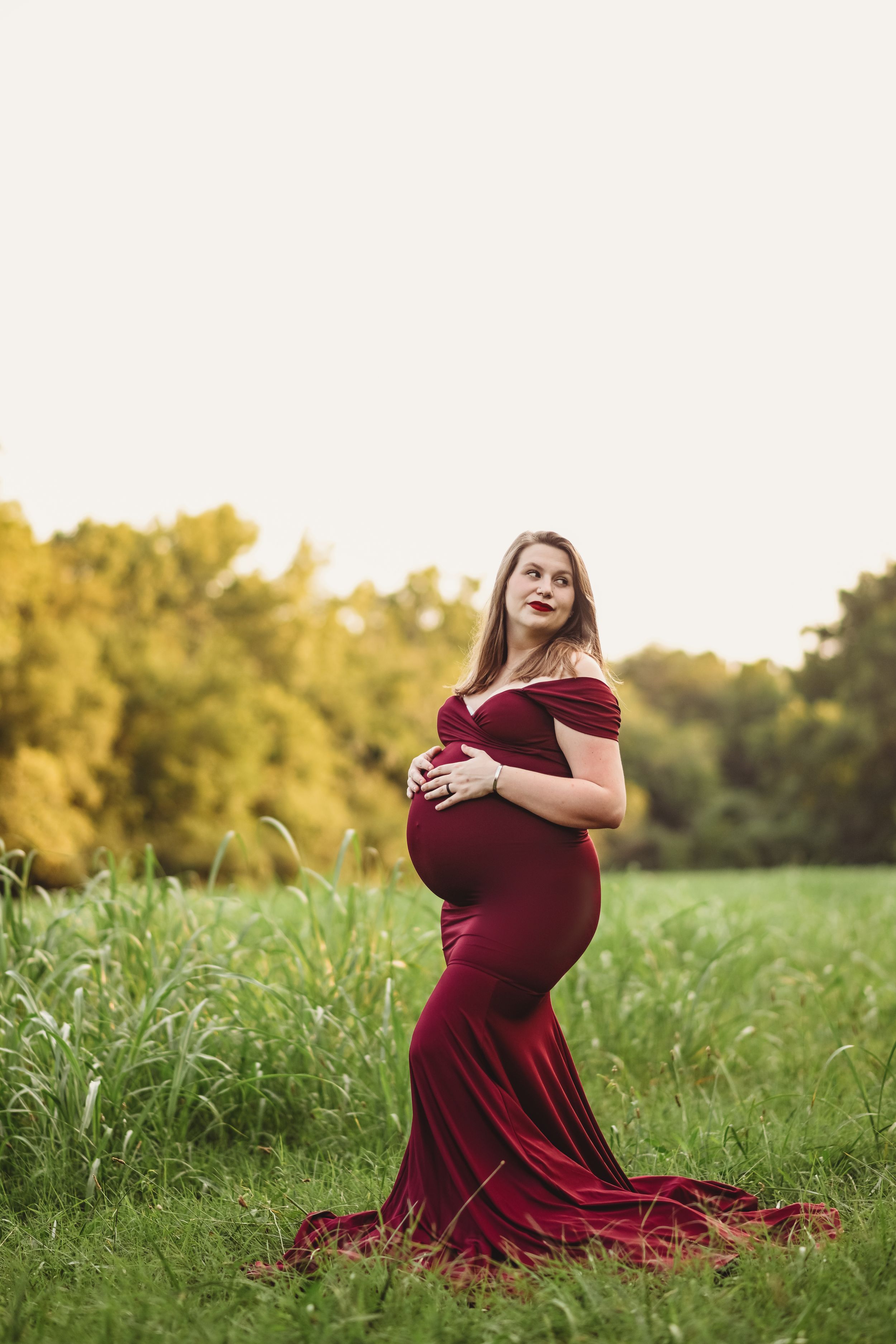 A maternity portrait of a woman in a burgundy dress standing in a sunlit field.