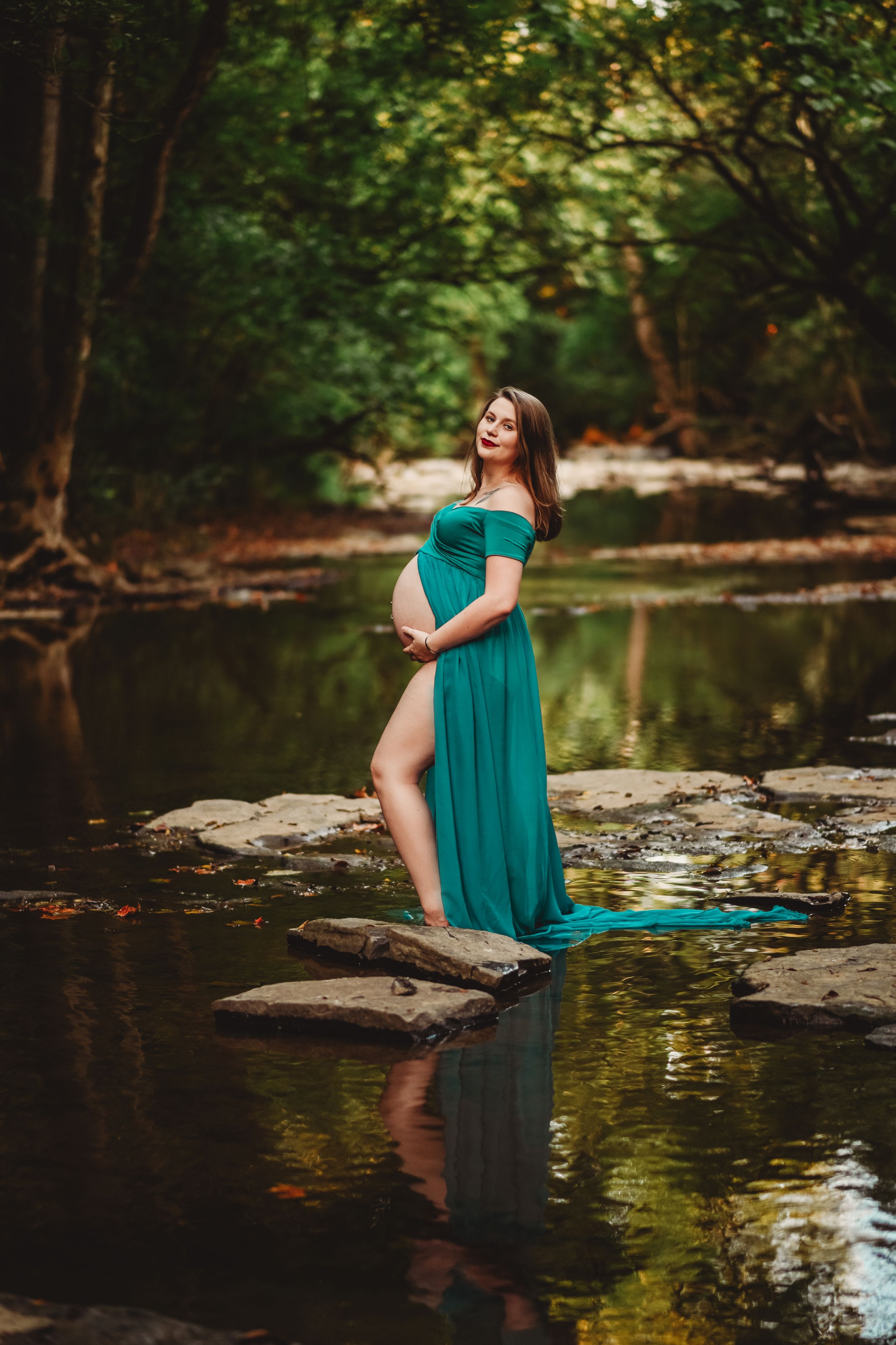 A teal dress flows elegantly by a tranquil pond with rocky edges in a wooded setting.