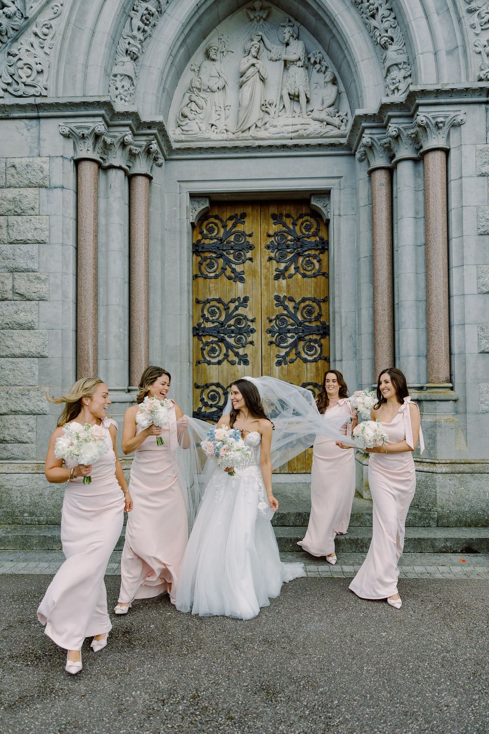 Bride in white gown with flowing veil walks with four bridesmaids in blush pink dresses outside ornate stone church.