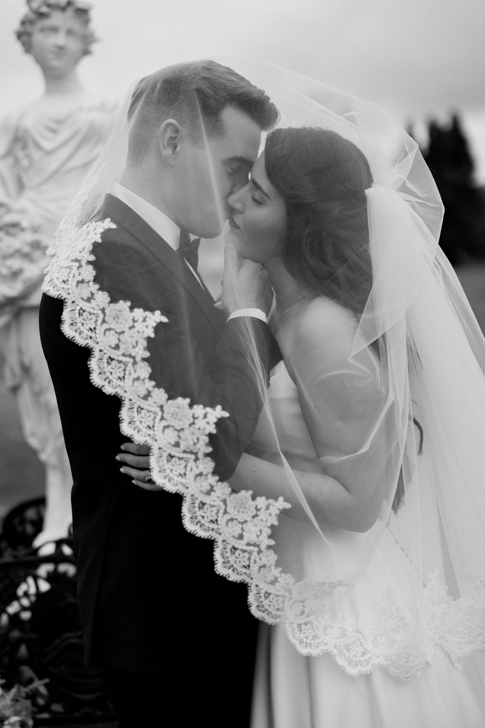 Black and white photo of a bride and groom sharing a kiss under a lace-trimmed veil near a stone statue.