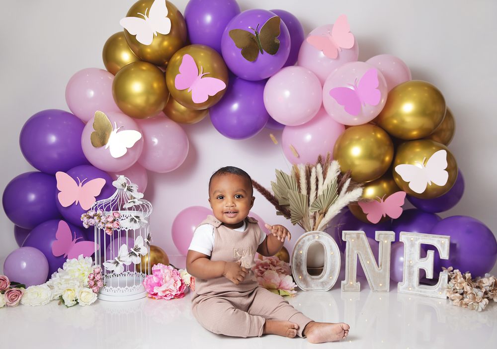 Baby in beige outfit sits by balloon arch with gold, purple and pink balloons decorated with butterflies and ONE letters.