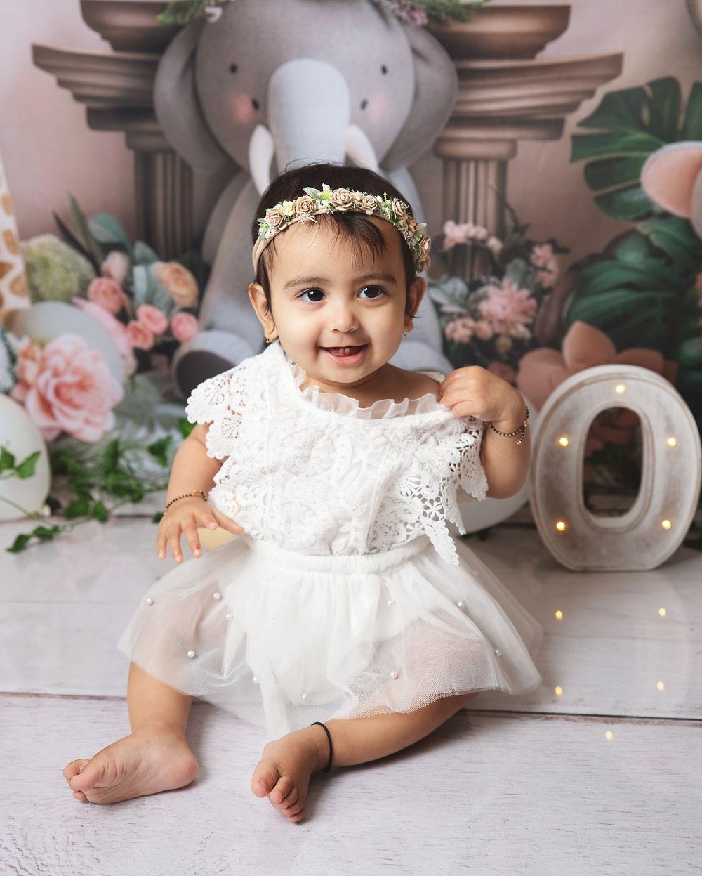 Sweet first birthday setup with floral decorations, plush elephant, and white tulle dress against a pink backdrop.