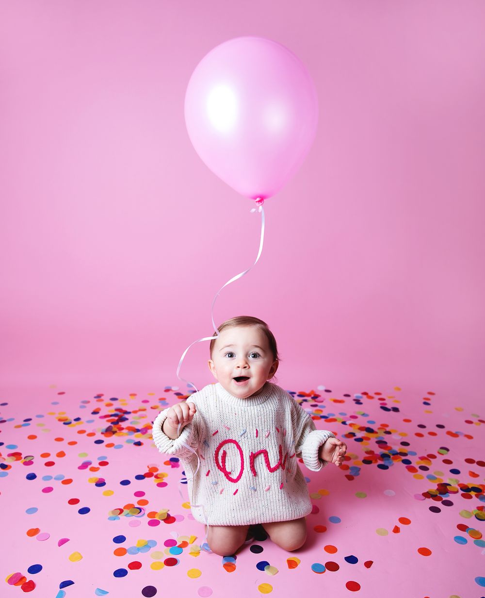 Small baby wearing 'one' sweater sits on pink backdrop with confetti scattered around and pink balloon floating above.