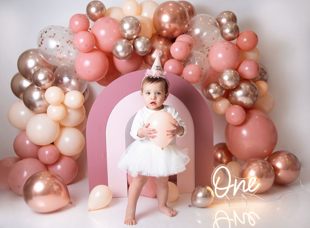 Elegant balloon arch decoration in pink, rose gold, and champagne colors with a pink backdrop and white text reading 'One'.