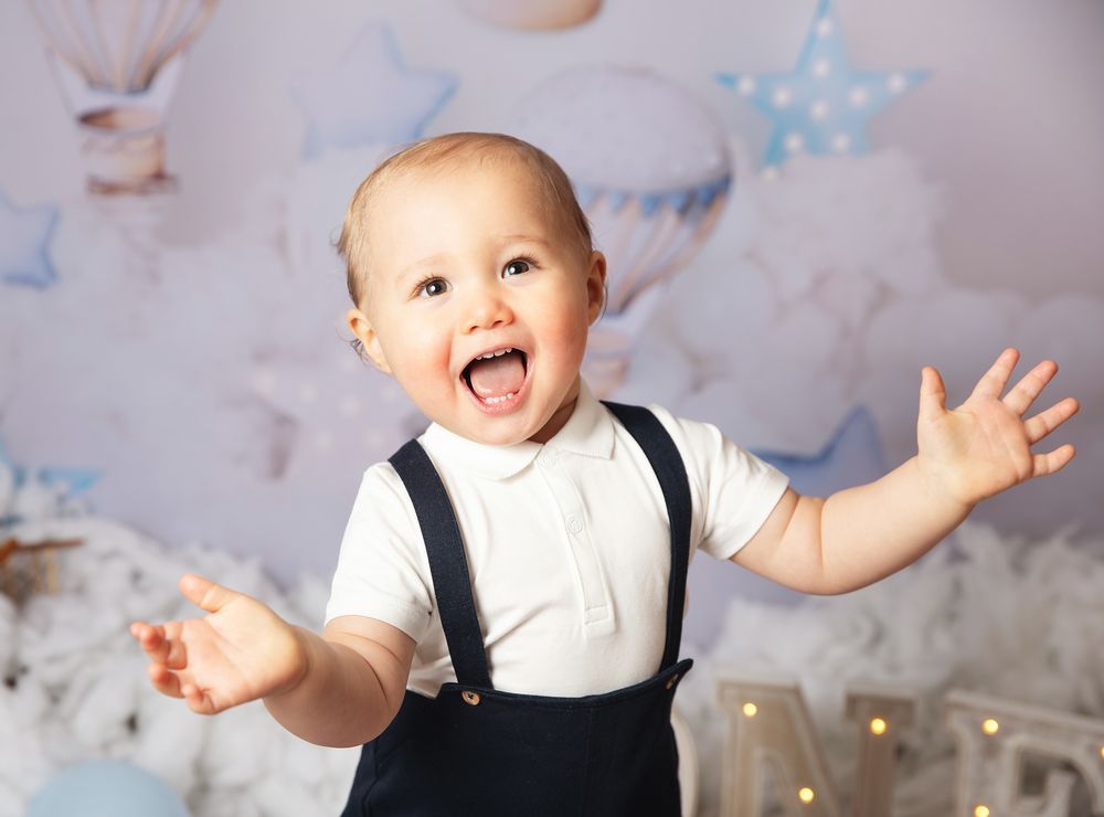 A joyful baby wearing black suspenders raises their arms in an expressive pose against a soft pastel background.